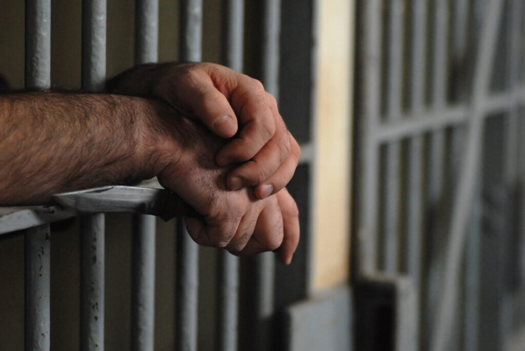 A person 's hands are folded over the bars of a jail cell.